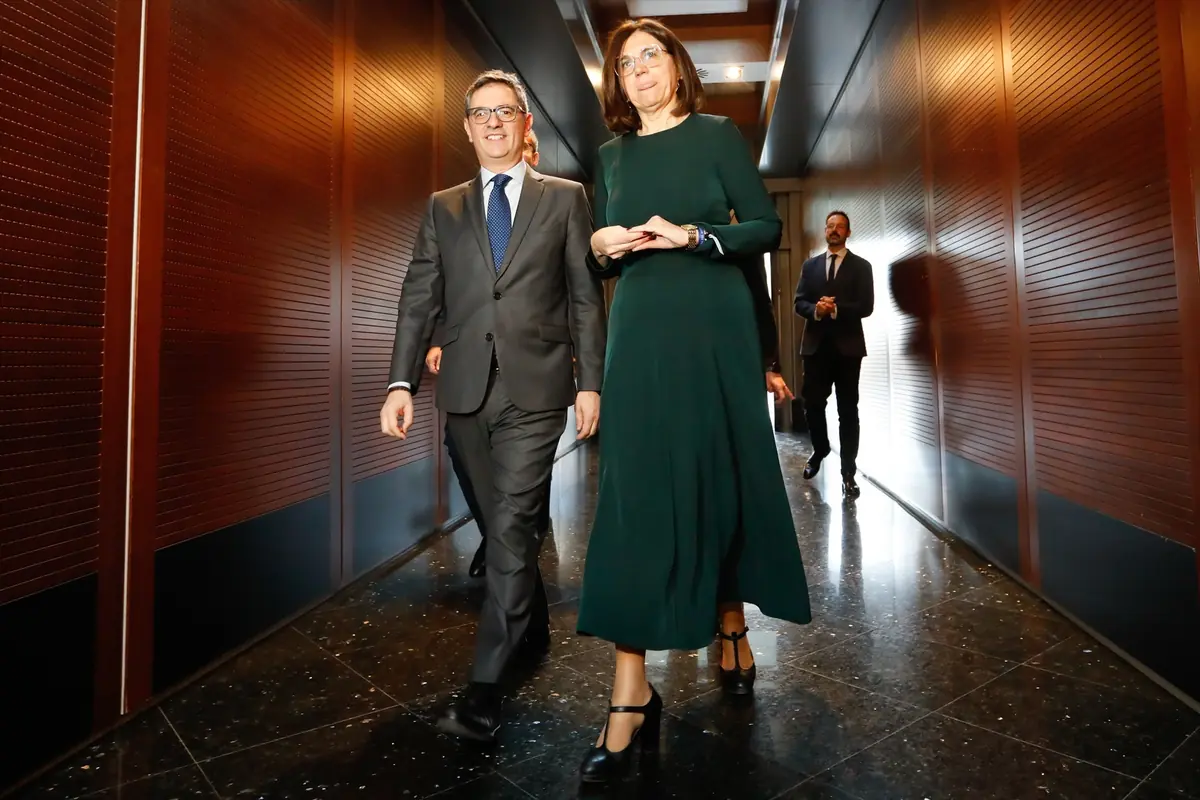 Dos personas con ropa formal caminando por un pasillo de oficina moderno con paredes de madera y suelo brillante