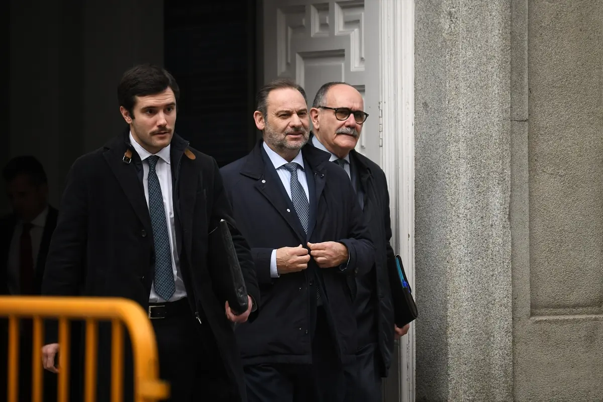 Tres hombres con traje y abrigo oscuro salen de un edificio oficial con carpetas en la mano mientras caminan junto a una valla amarilla