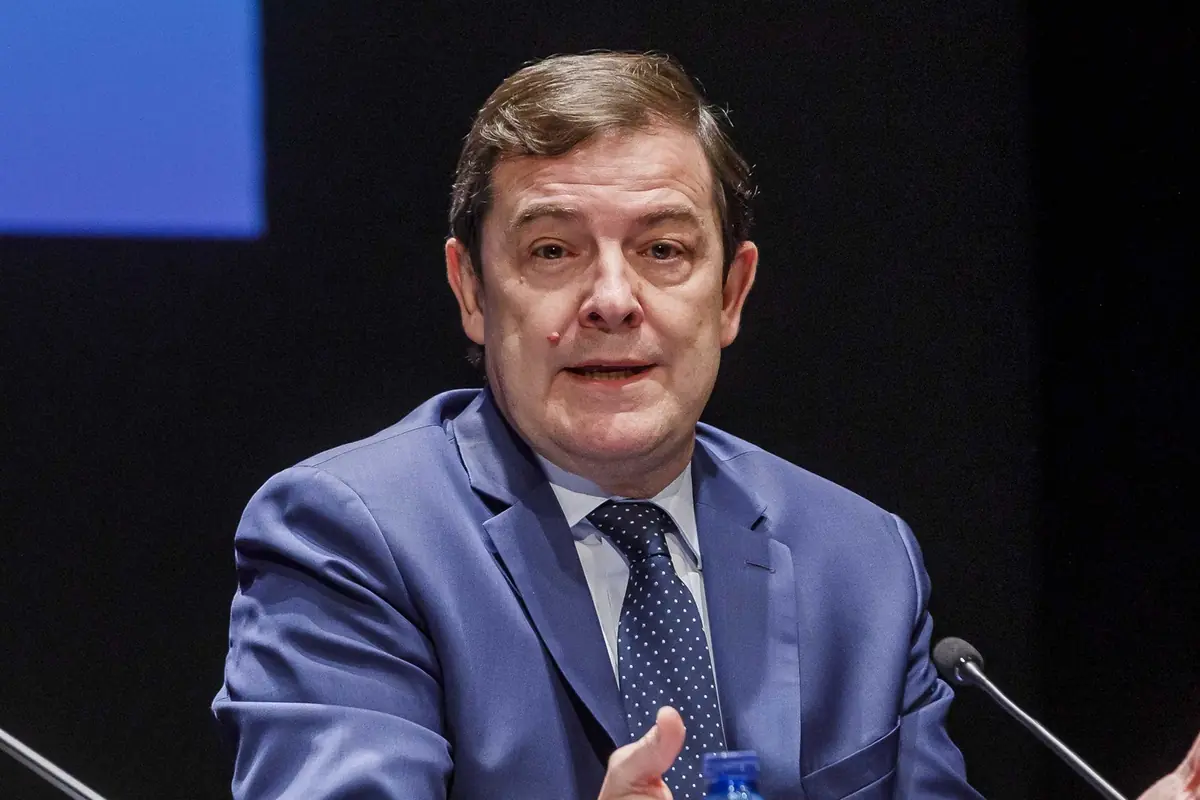Hombre de mediana edad con traje azul hablando en una conferencia sentado frente a micrófonos y una botella de agua