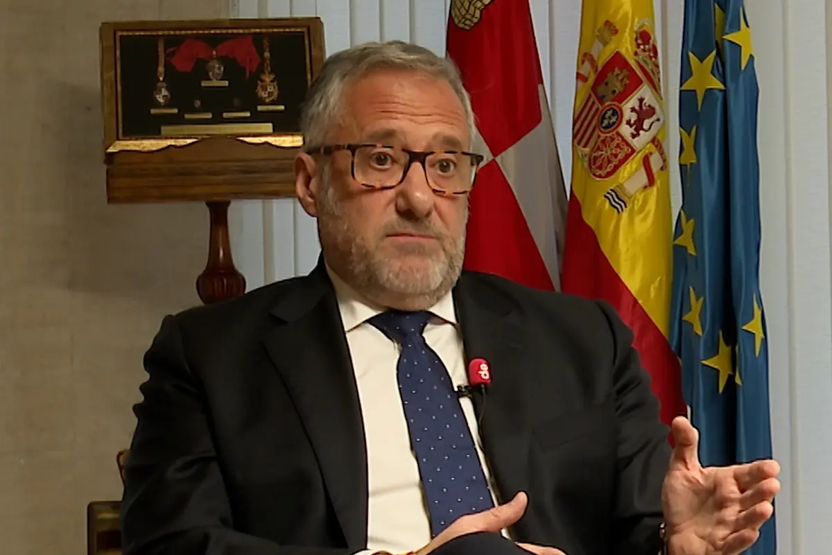 Hombre de mediana edad con traje oscuro y corbata azul hablando en una entrevista en un despacho con banderas de España y de la Unión Europea al fondo