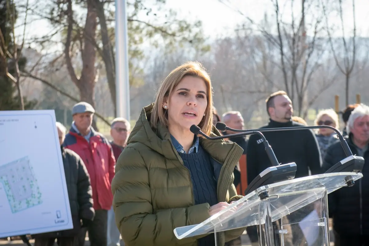 Mujer con abrigo verde hablando en un atril transparente al aire libre frente a un grupo de personas