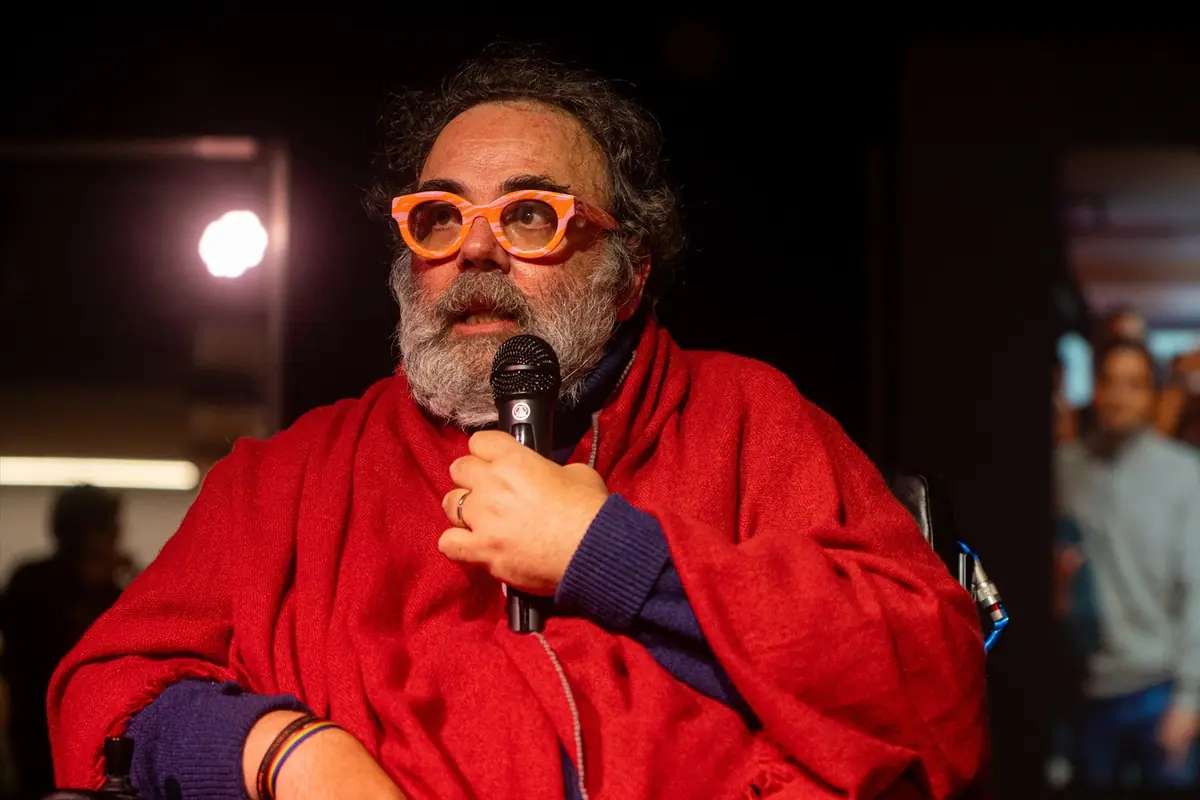 Hombre de mediana edad con barba canosa y gafas naranjas hablando por un micrófono mientras viste una manta roja en un escenario oscuro Hombre de mediana edad con barba canosa y gafas naranjas hablando por un micrófono mientras viste una manta roja en un escenario oscuro