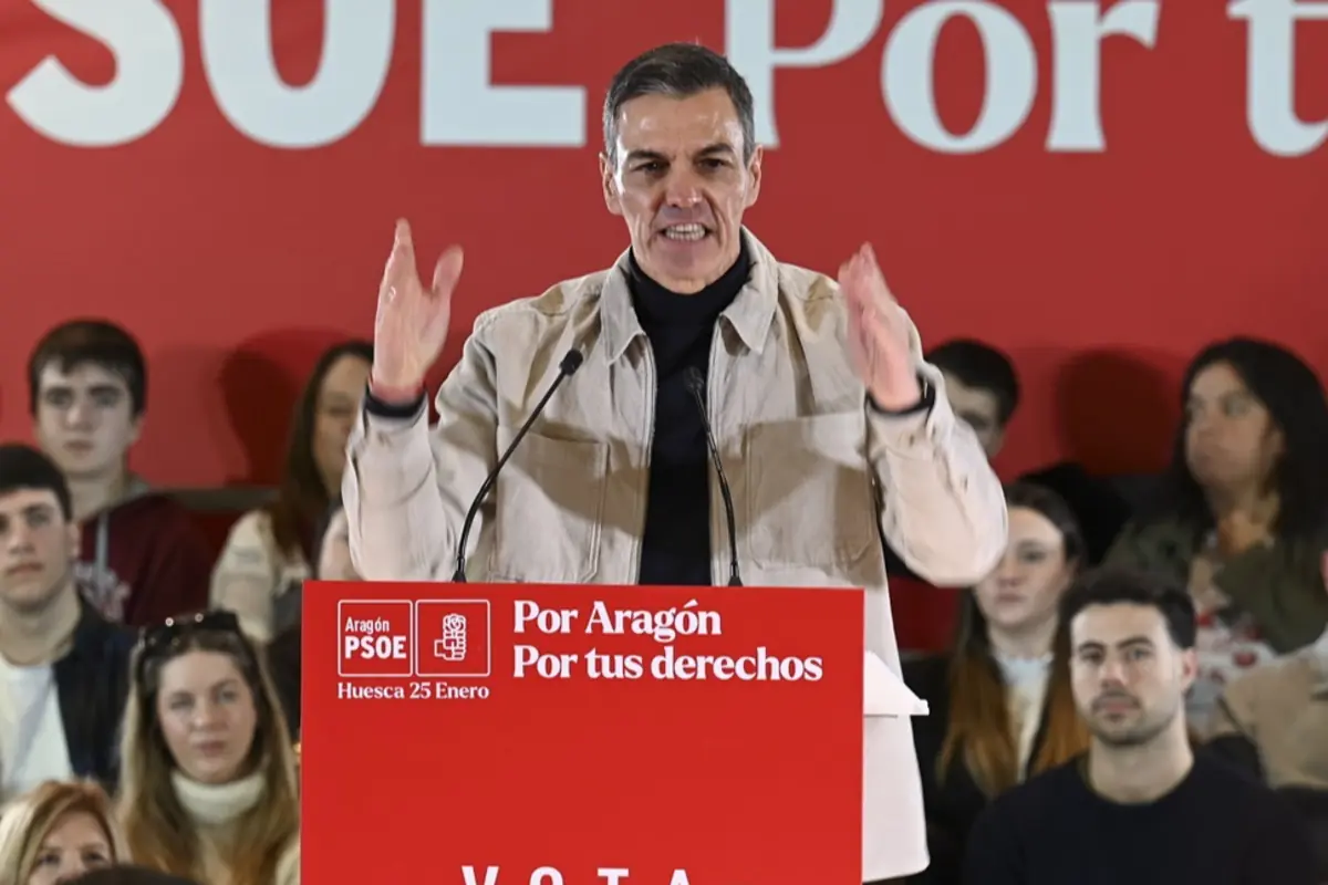 Hombre hablando en un mitin político del PSOE en un escenario rojo frente a un atril con el lema Por Aragón Por tus derechos rodeado de público