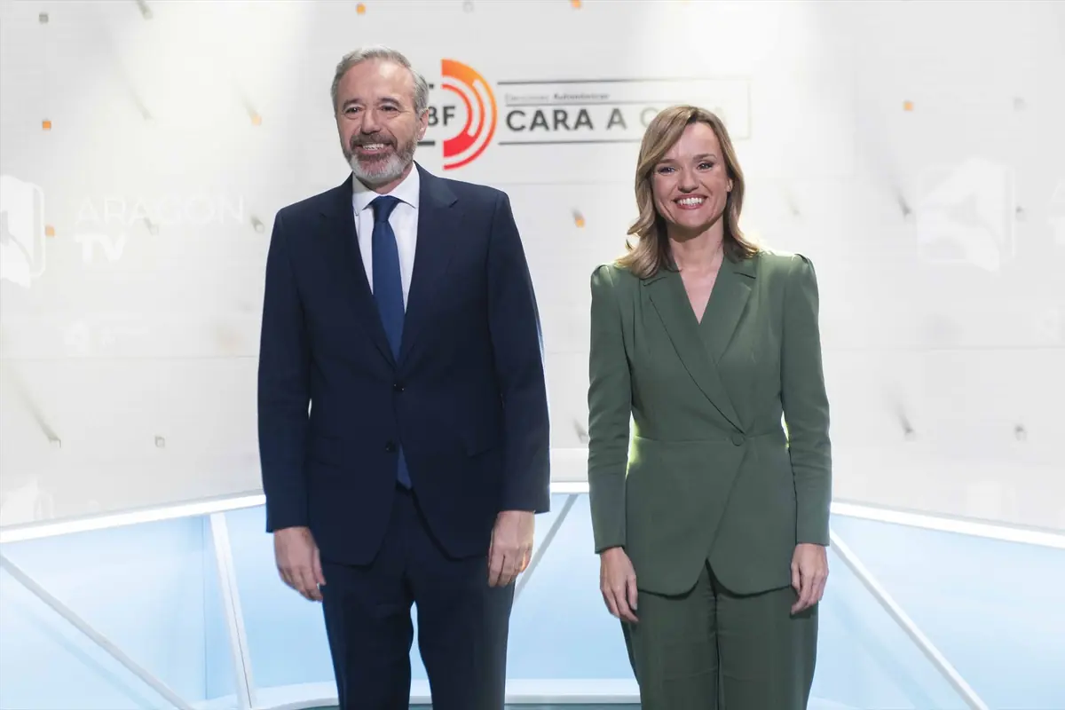 Dos presentadores de televisión de pie en un plató luminoso con el logotipo de Aragón TV al fondo Dos presentadores de televisión de pie en un plató luminoso con el logotipo de Aragón TV al fondo