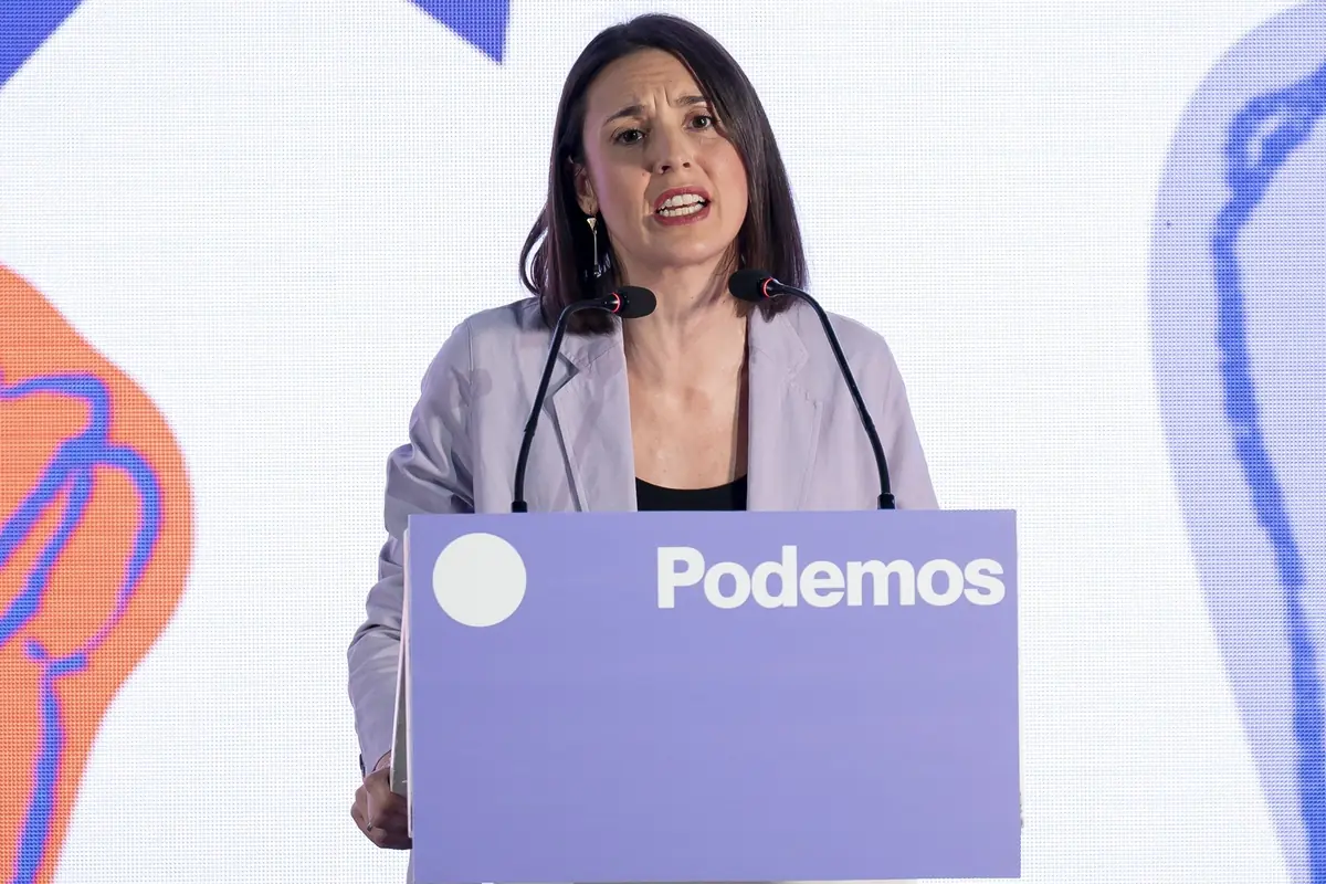 Mujer hablando en un atril morado con el logotipo y la palabra Podemos durante un acto público