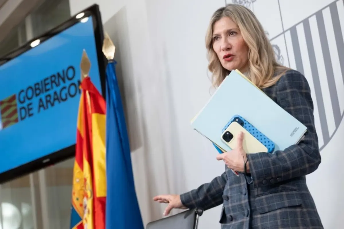 Mujer rubia con carpeta y móvil en la mano durante una comparecencia junto a banderas y una pantalla con el texto Gobierno de Aragón