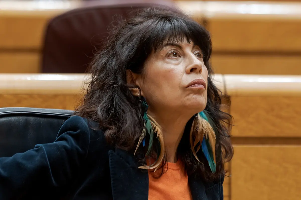 Mujer de mediana edad con pendientes largos de plumas sentada en un recinto interior mirando hacia arriba con expresión seria