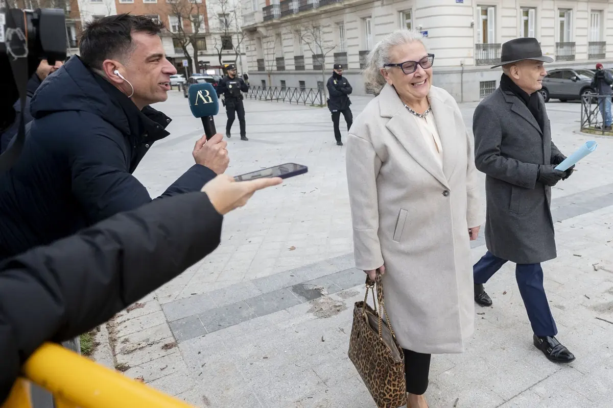 Periodista con micrófono entrevista en la calle a una mujer mayor sonriente que camina con abrigo claro y bolso de estampado animal acompañada por un hombre con sombrero y abrigo oscuro mientras policías vigilan al fondo