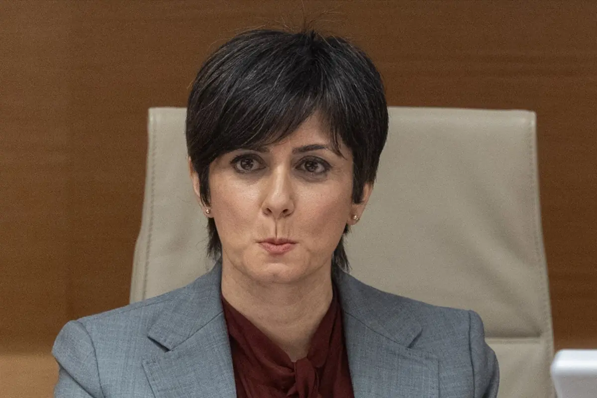 Mujer de mediana edad con pelo corto oscuro y traje gris sentada en una silla clara mirando al frente con gesto serio