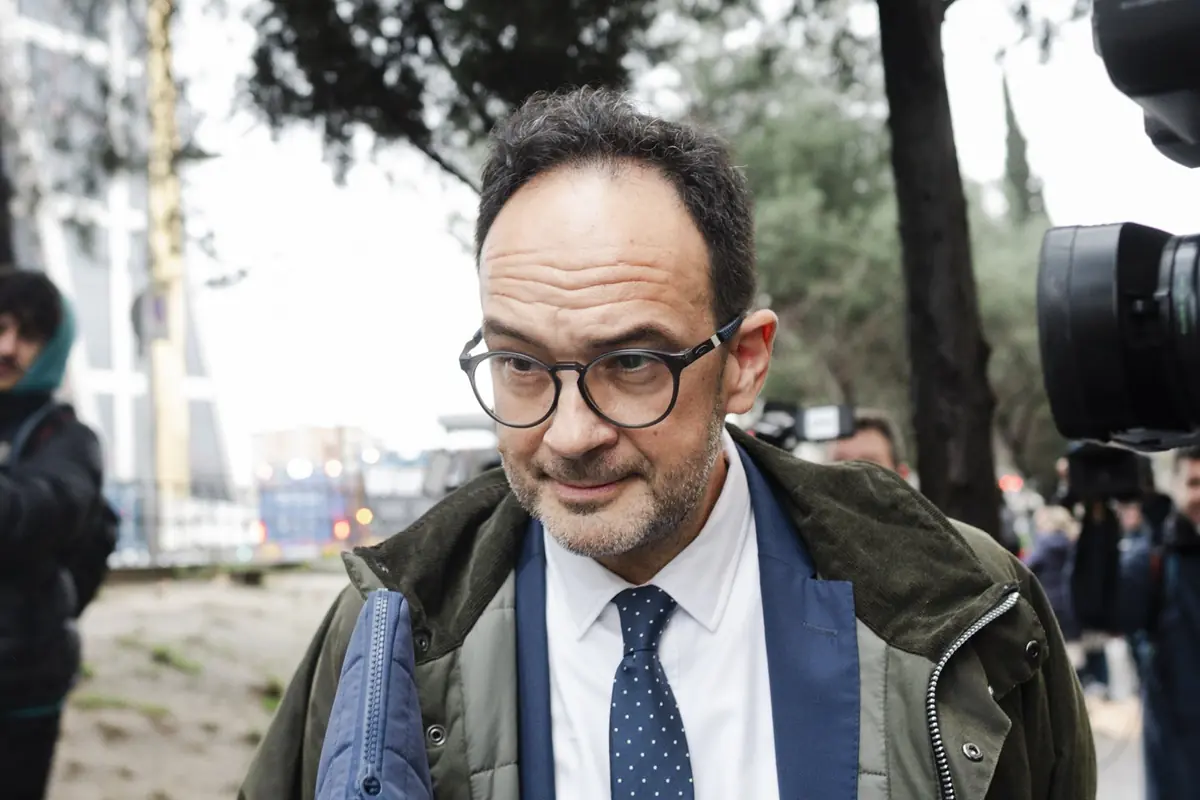 Hombre de mediana edad con gafas y traje caminando al aire libre rodeado de cámaras y periodistas