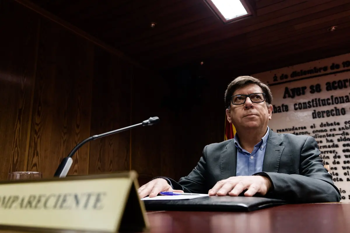 Hombre con gafas y traje sentado ante un micrófono en una sala de conferencias con un cartel de texto grande al fondo Hombre con gafas y traje sentado ante un micrófono en una sala de conferencias con un cartel de texto grande al fondo