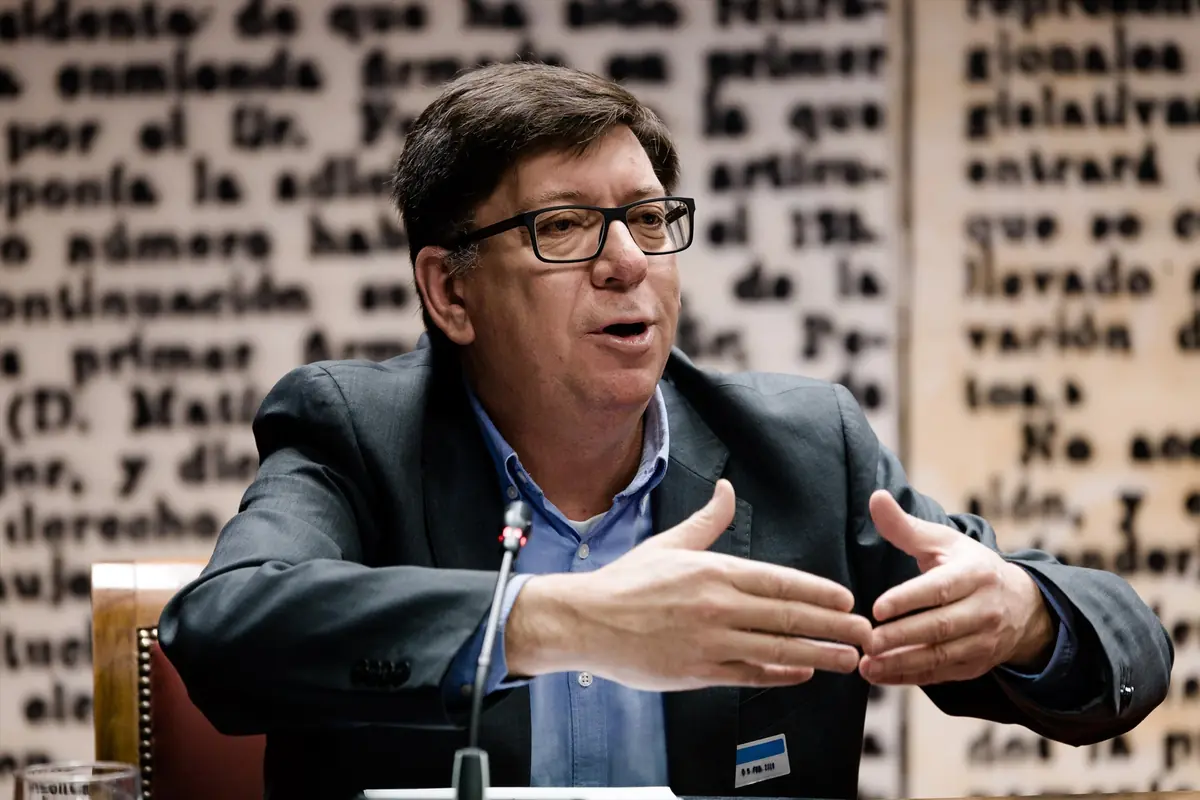 Hombre de mediana edad con gafas y traje hablando gesticulando con las manos frente a un micrófono y un fondo de texto borroso