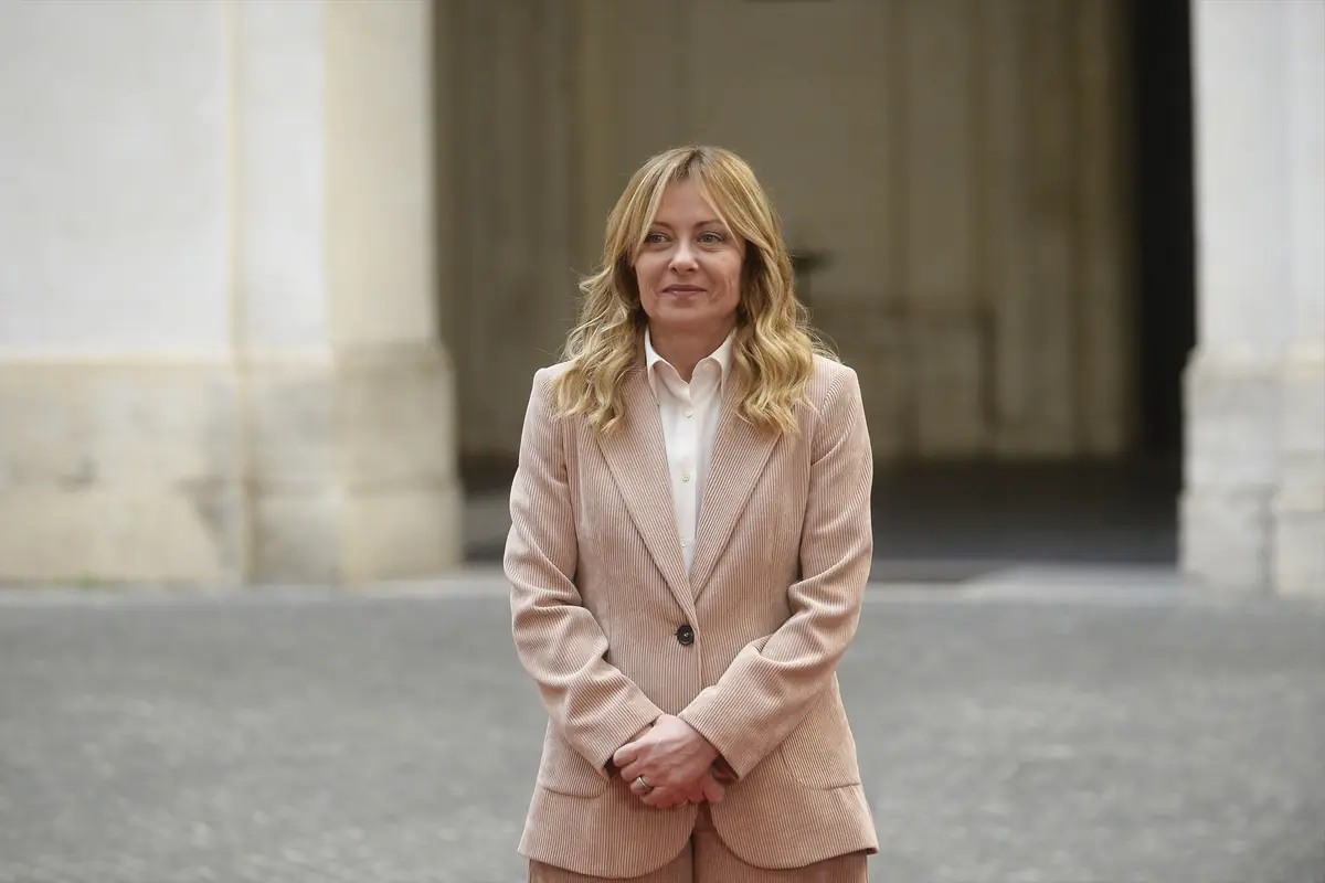 Mujer rubia de pie al aire libre con traje de chaqueta beige y camisa blanca frente a un edificio de piedra
