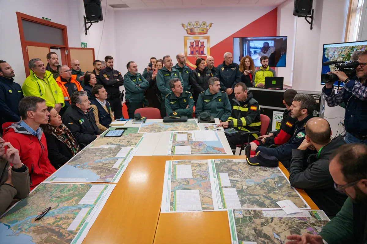 Reunión de coordinación de equipos de emergencia y fuerzas de seguridad alrededor de una mesa llena de mapas y documentos en una sala de mando equipada con pantallas y cámaras