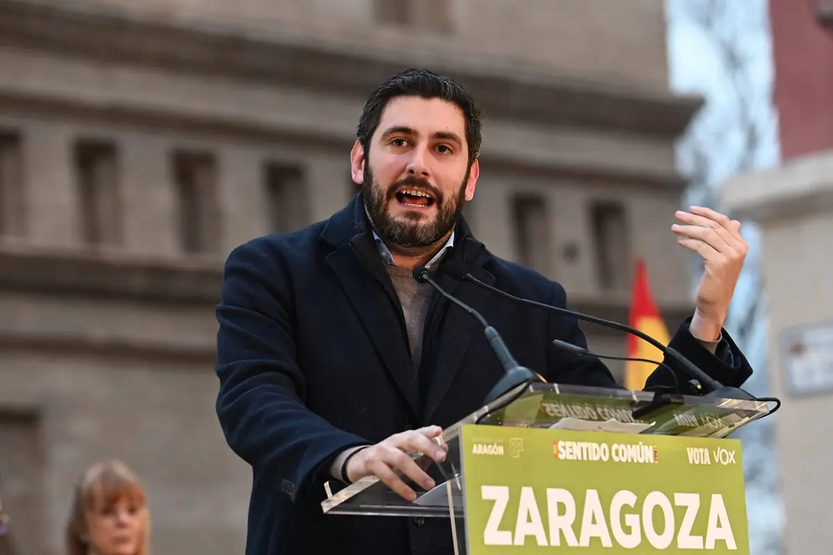 Hombre hablando en un mitin político al aire libre en Zaragoza detrás de un atril con micrófonos y un cartel verde de campaña