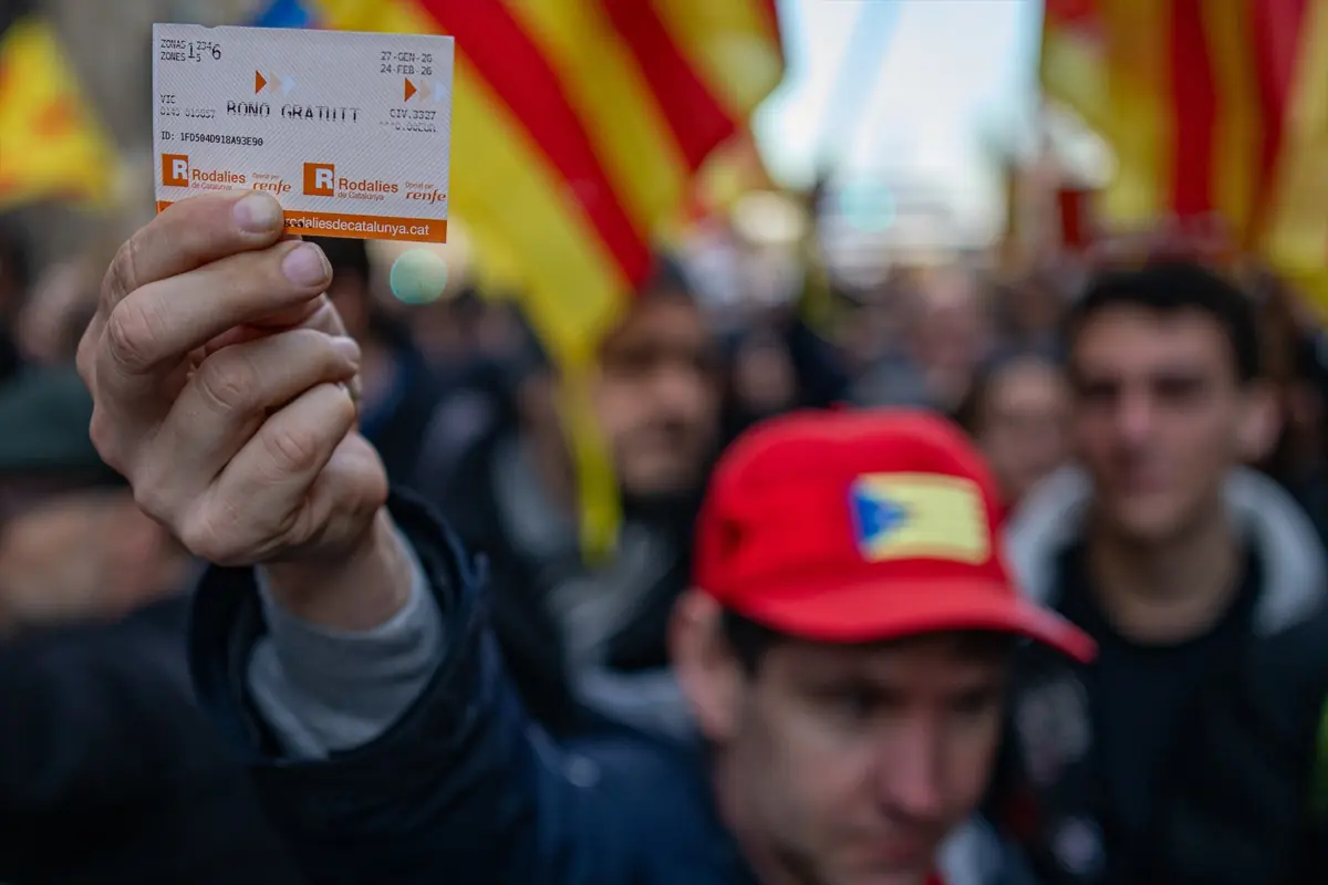 Persona en una manifestación sostiene en primer plano un billete de Rodalies de Catalunya mientras al fondo se ve una multitud con banderas catalanas