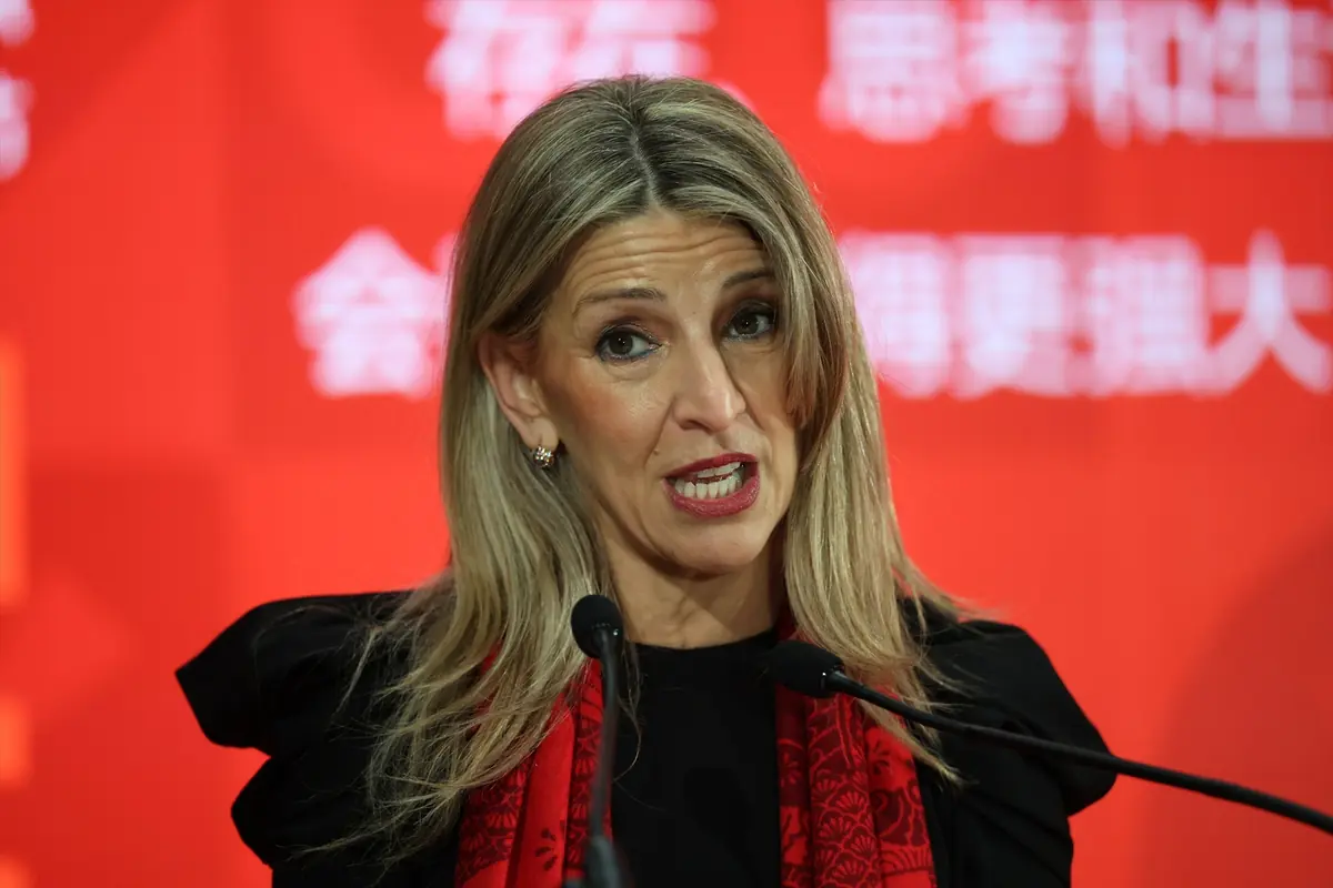 Mujer rubia hablando frente a micrófonos con fondo rojo y bufanda roja