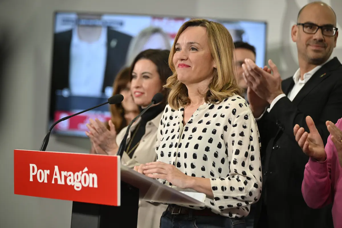 Mujer sonriente dando un discurso en un atril rojo con el texto Por Aragón rodeada de personas que aplauden en un acto político