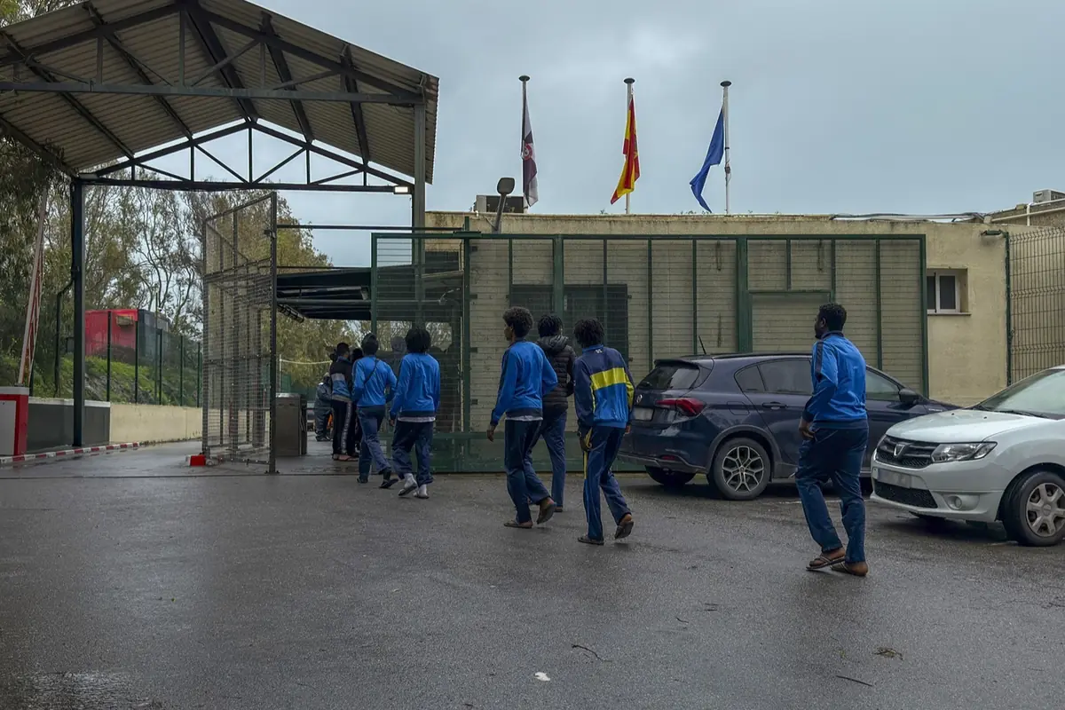 Grupo de personas con ropa deportiva azul caminando hacia la entrada de un recinto vallado con coches aparcados y banderas ondeando sobre el edificio