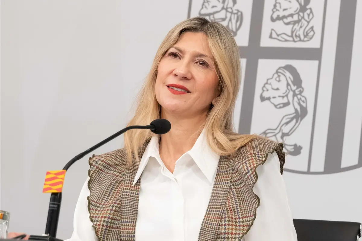 Mujer rubia hablando frente a un micrófono en una rueda de prensa con fondo institucional