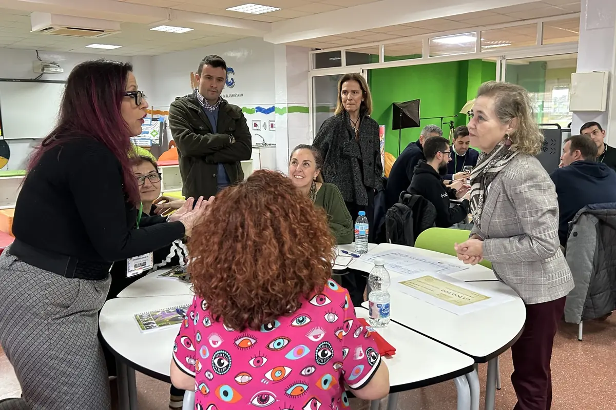 Varias personas dialogan y colaboran alrededor de una mesa en un aula moderna con paredes verdes y material de trabajo sobre la superficie