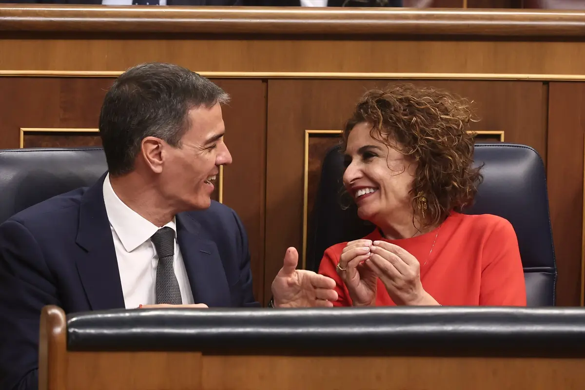 Dos personas sentadas en un escaño parlamentario conversan y sonríen entre sí mientras gesticulan con las manos Dos personas sentadas en un escaño parlamentario conversan y sonríen entre sí mientras gesticulan con las manos