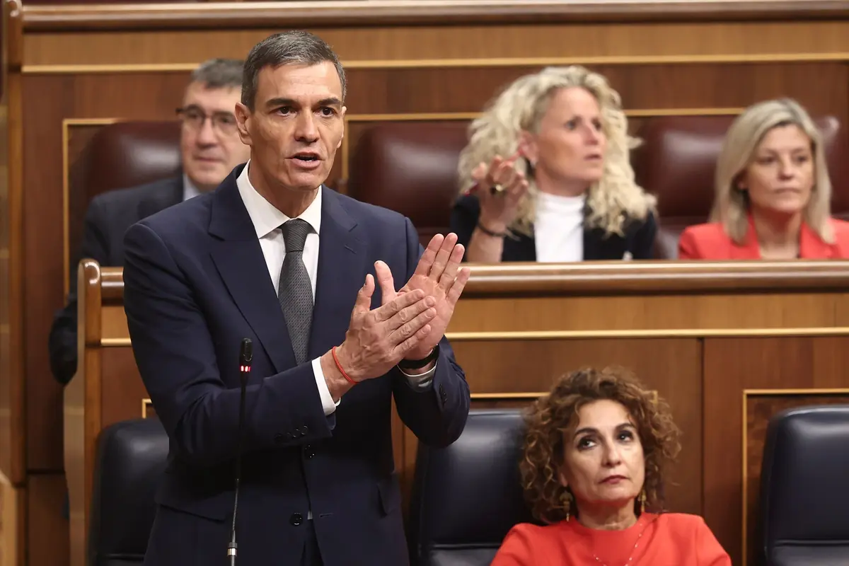 Hombre con traje oscuro hablando y gesticulando de pie en un parlamento con otras personas sentadas detrás en sus escaños