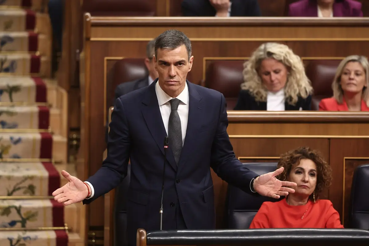 Hombre con traje oscuro hablando con gesto serio y brazos abiertos en un parlamento lleno de asistentes
