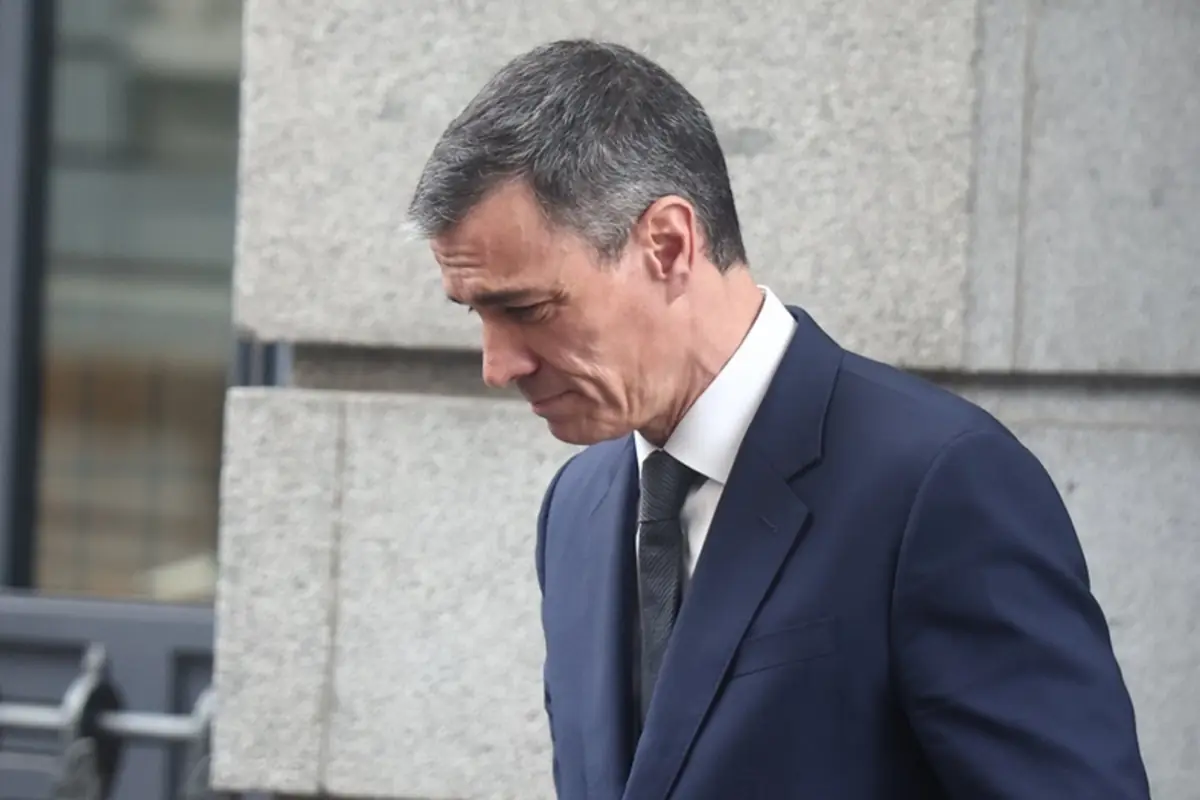 Hombre de mediana edad con traje azul marino y corbata oscura caminando cabizbajo junto a una pared de piedra gris