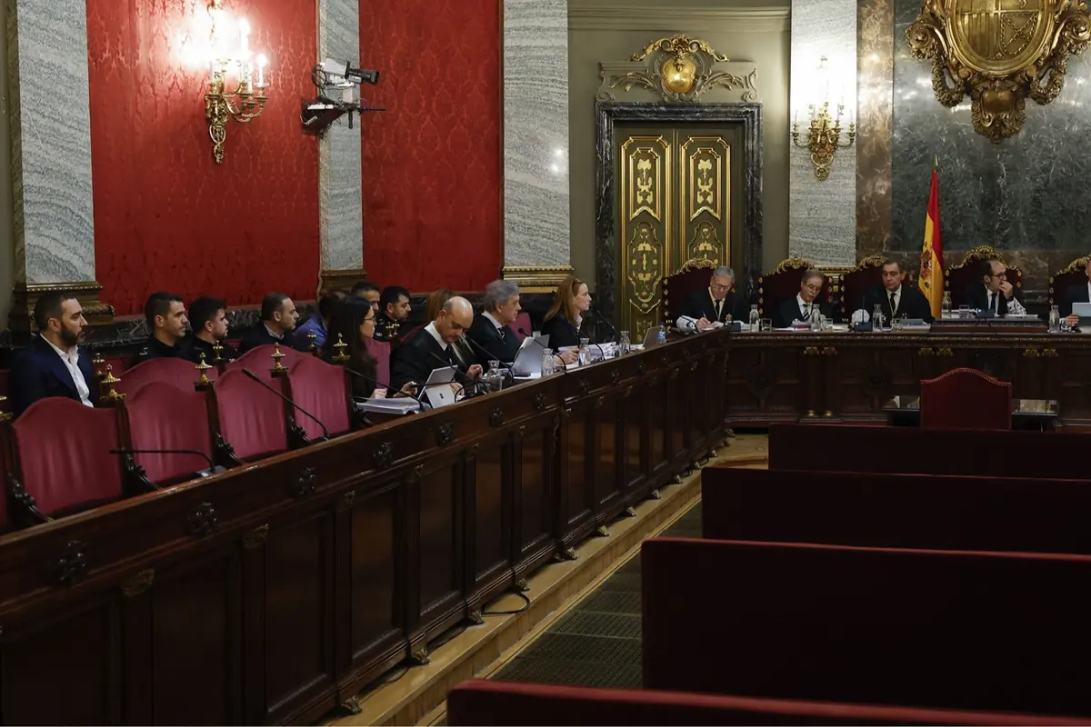 Sala de tribunal con jueces y abogados sentados en una mesa larga en un salón solemne con paredes rojas y la bandera de España al fondo