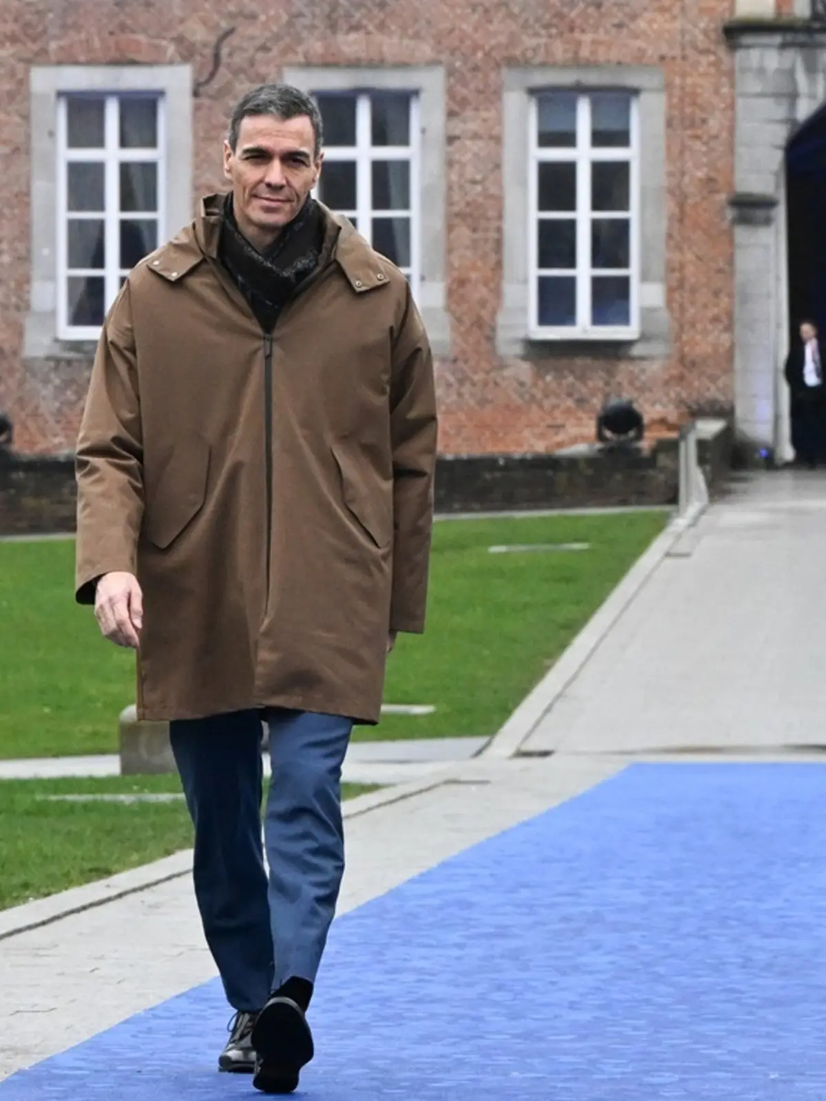 Hombre con abrigo marrón caminando sobre una alfombra azul en el exterior de un edificio de ladrillo con ventanas blancas