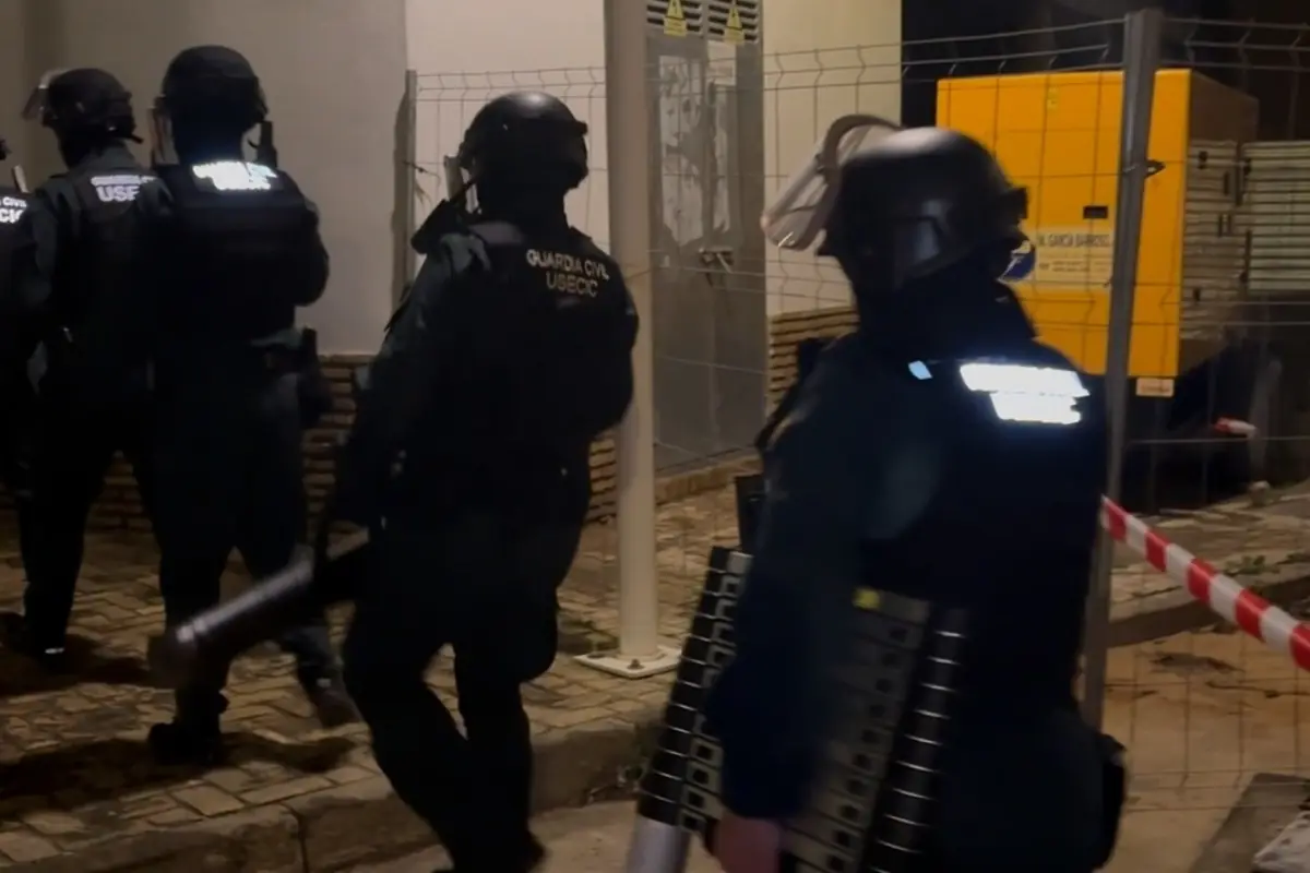 Agentes antidisturbios de la Guardia Civil equipados con cascos y protecciones avanzan de noche por una zona acordonada con vallas metálicas y cinta roja y blanca