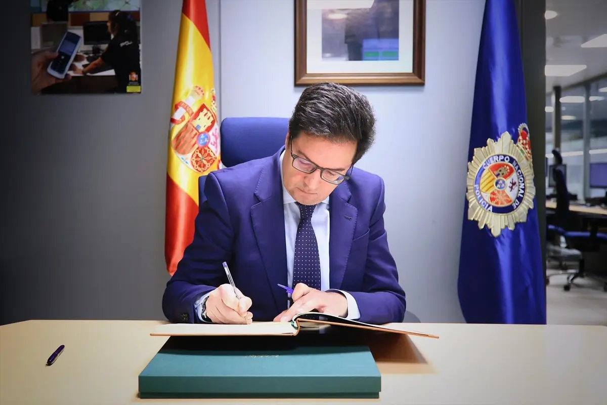 Hombre con traje azul firmando un documento en una oficina oficial entre la bandera de España y un estandarte del cuerpo de policía