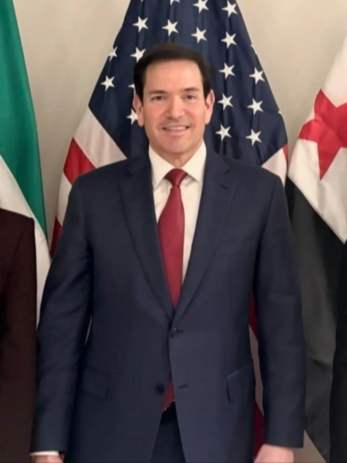 Hombre con traje azul y corbata roja posando frente a una bandera de Estados Unidos