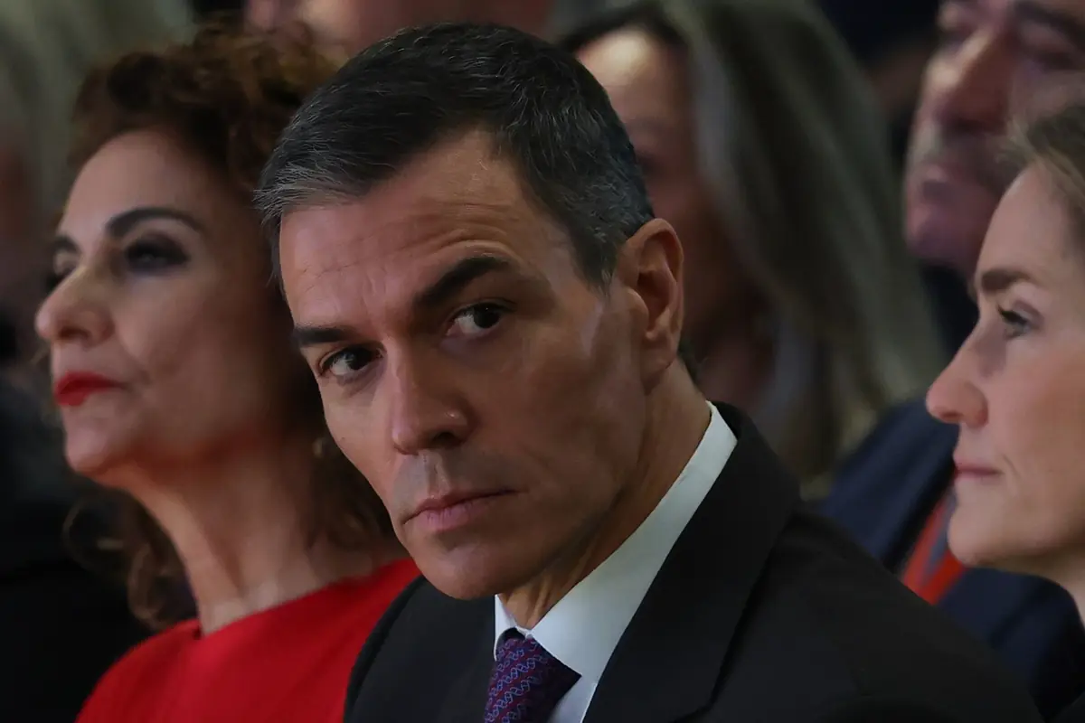 Hombre de mediana edad con traje oscuro y corbata sentado entre otras personas en un acto público mirando con expresión seria hacia un lado