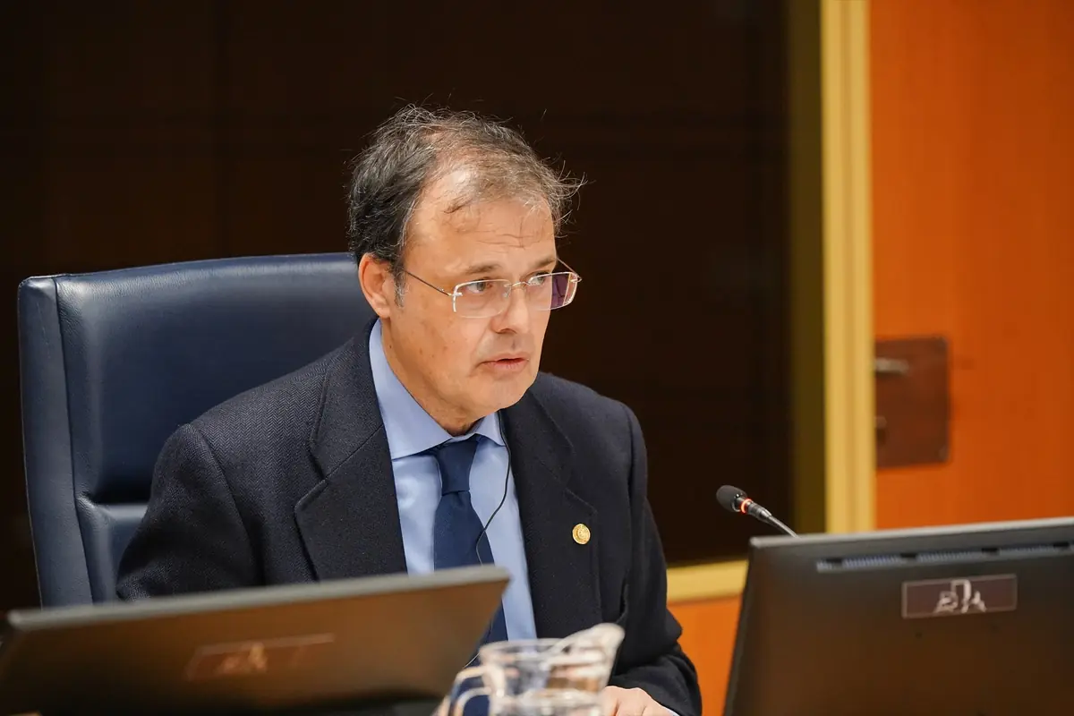 Hombre de mediana edad con traje oscuro y corbata hablando frente a un micrófono en una sala de reuniones oficiales