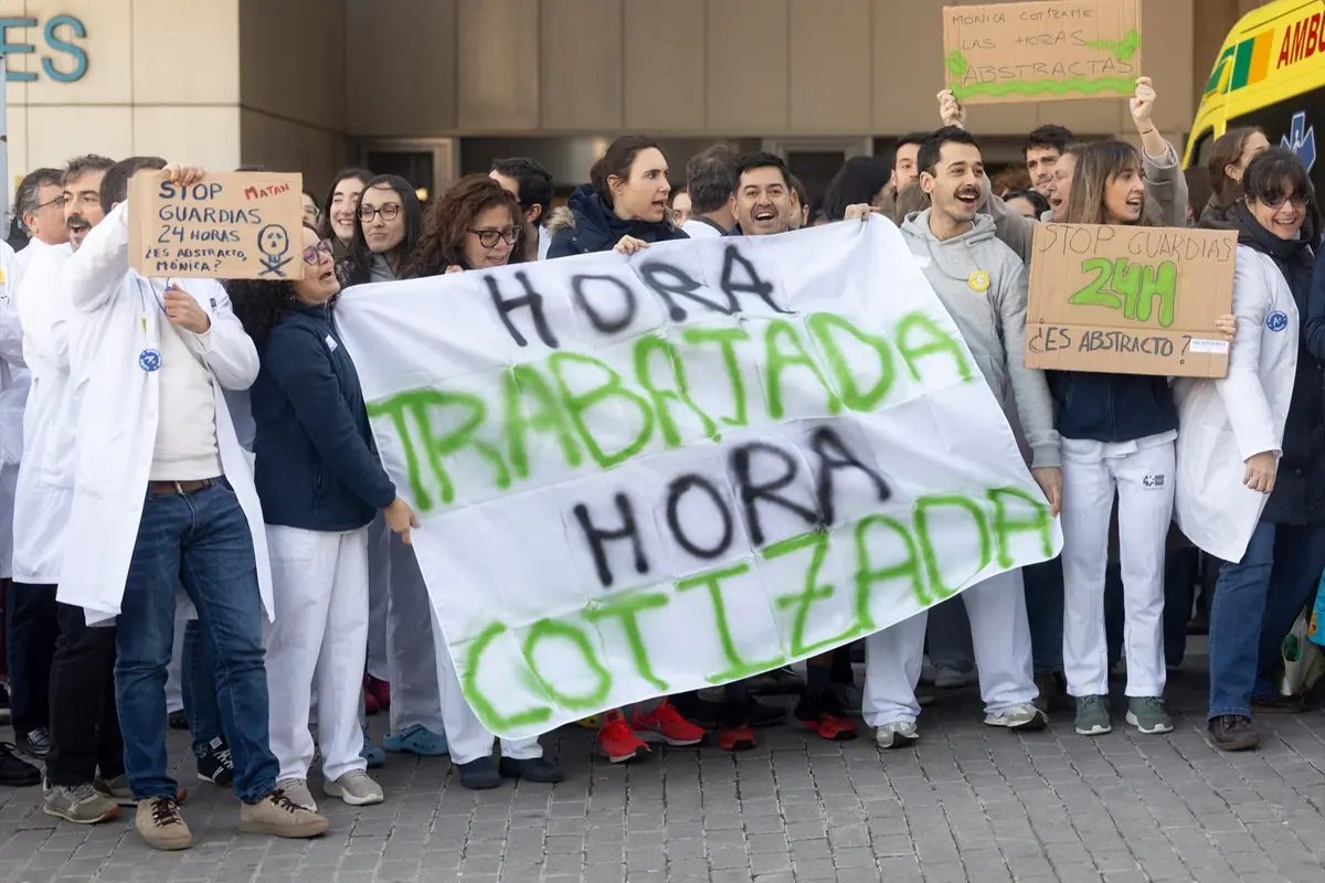 Grupo de personal sanitario con batas blancas y ropa médica protesta en la calle frente a un edificio sosteniendo pancartas y una gran sábana donde se lee hora trabajada hora cotizada
