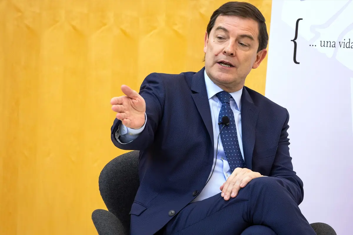 Hombre con traje azul hablando en una conferencia y gesticulando con la mano extendida mientras está sentado en una silla