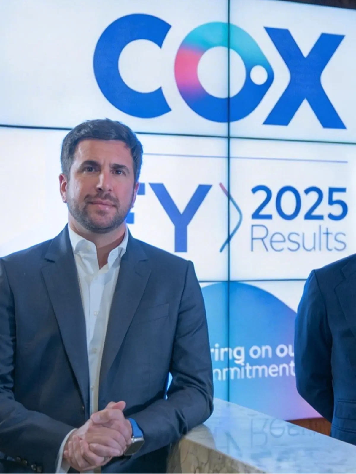 Hombre con traje oscuro posando frente a una pantalla que muestra el logotipo de COX y resultados financieros para 2025 Hombre con traje oscuro posando frente a una pantalla que muestra el logotipo de COX y resultados financieros para 2025