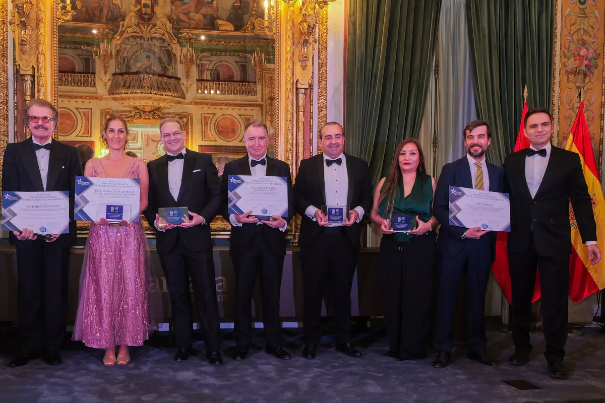 Grupo de personas elegantemente vestidas posando en un salón lujoso mientras sostienen diplomas y trofeos de un acto de premiación