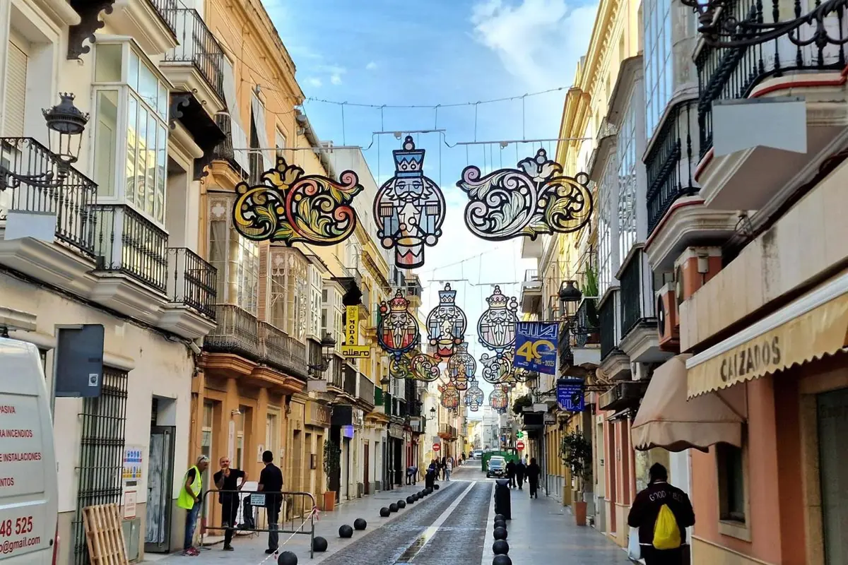 Calle peatonal de una ciudad española con edificios antiguos y balcones, decorada con luces festivas colgantes con figuras de los Reyes Magos y algunas personas caminando y trabajando en la vía Calle peatonal de una ciudad española con edificios antiguos y balcones, decorada con luces festivas colgantes con figuras de los Reyes Magos y algunas personas caminando y trabajando en la vía