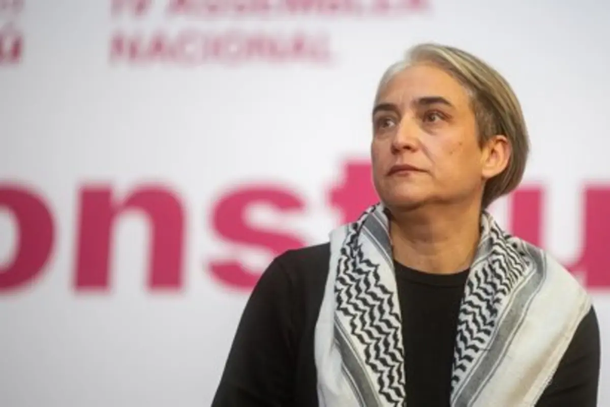Mujer de mediana edad con pañuelo blanco y negro al cuello mirando hacia un lado frente a un fondo con letras rojas desenfocadas