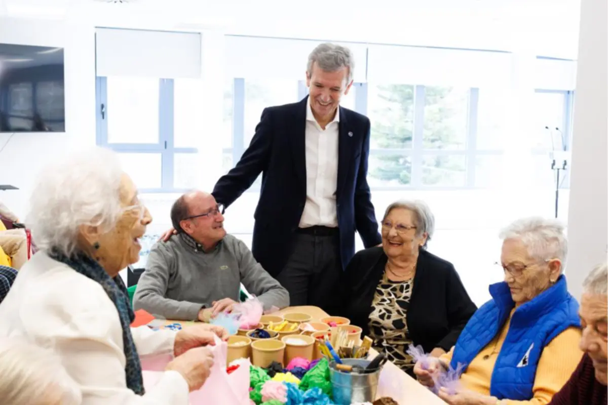Grupo de personas mayores realizando manualidades en una mesa mientras un hombre de traje las acompaña y conversa en un ambiente luminoso y alegre