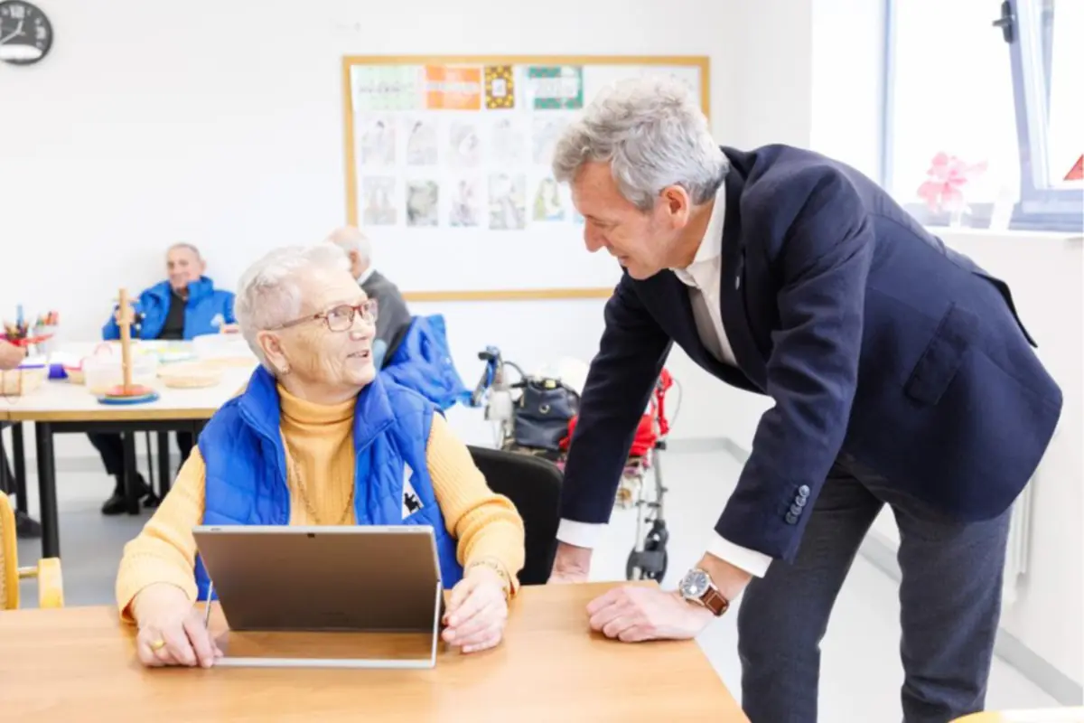 Hombre de mediana edad conversa sonriente con una mujer mayor que usa una tablet en una sala luminosa de centro de día con otras personas al fondo Hombre de mediana edad conversa sonriente con una mujer mayor que usa una tablet en una sala luminosa de centro de día con otras personas al fondo