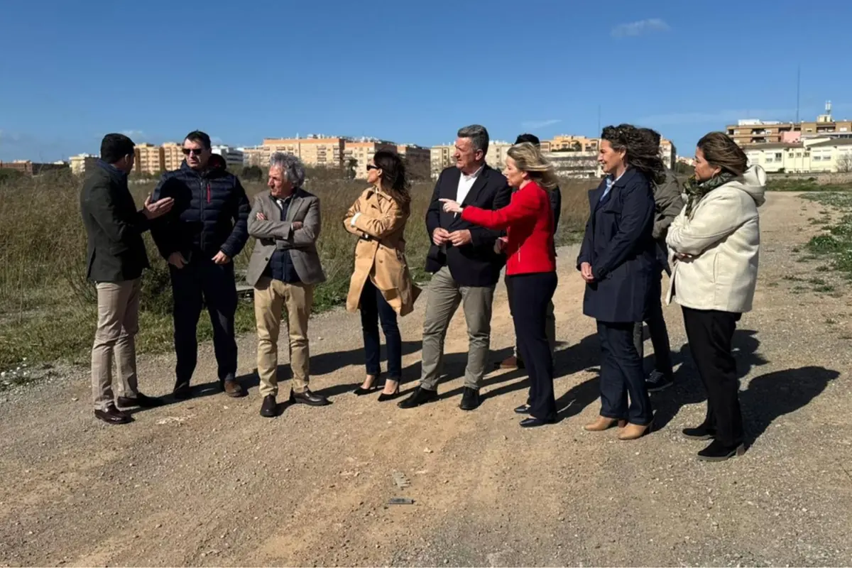 Grupo de personas de pie en un descampado soleado conversando y señalando el terreno con edificios residenciales al fondo