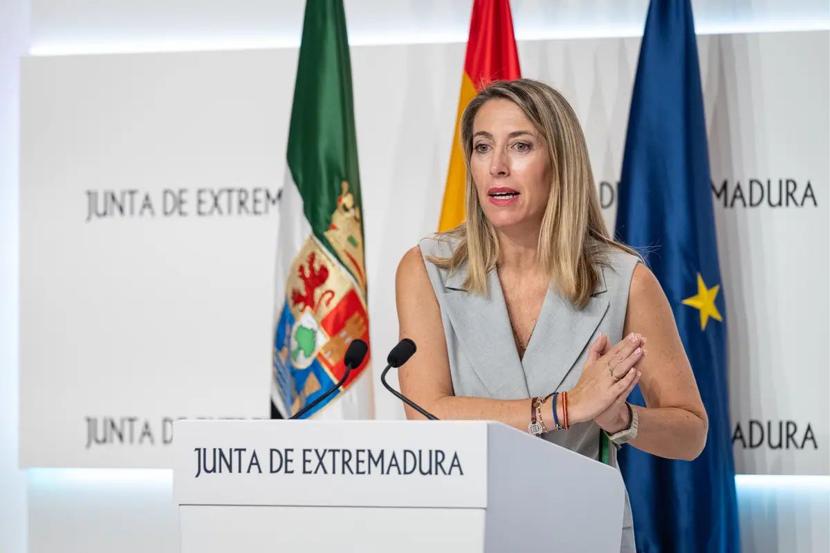 Mujer rubia hablando en una rueda de prensa en un atril de la Junta de Extremadura con banderas oficiales de fondo