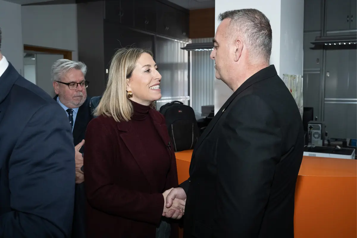 Dos personas vestidas de forma formal se dan la mano y conversan sonrientes en una oficina mientras otra persona observa al fondo