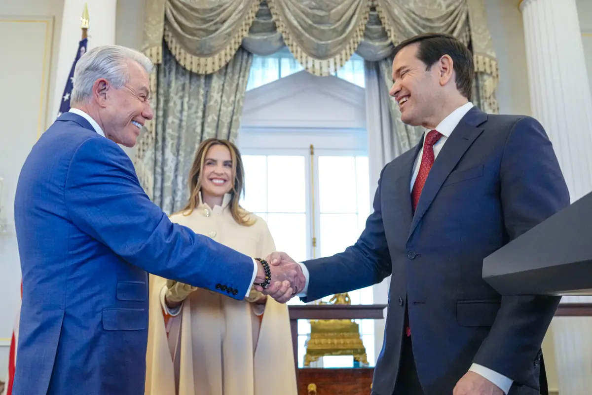 Dos hombres de traje se dan la mano sonrientes en un salón elegante mientras una mujer de pie detrás de ellos observa la escena con una sonrisa