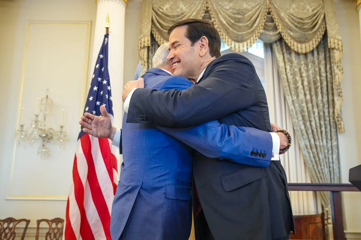 Dos hombres con traje se abrazan sonrientes en un salón oficial junto a una bandera de Estados Unidos