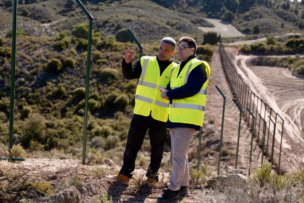 Dos trabajadores con chalecos reflectantes conversan y señalan el terreno junto a una valla en una zona rural montañosa Dos trabajadores con chalecos reflectantes conversan y señalan el terreno junto a una valla en una zona rural montañosa